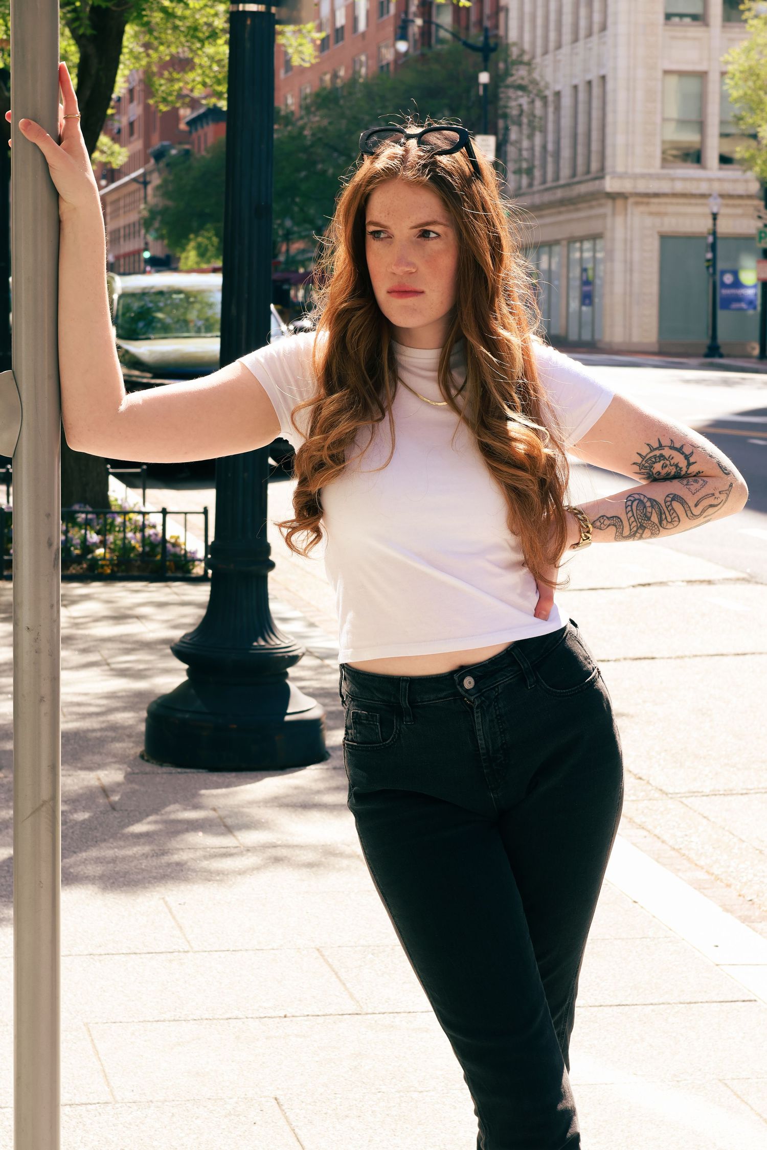 Young woman with long wavy red hair in white tee and black jeans, leaning on pole on sunny city street.