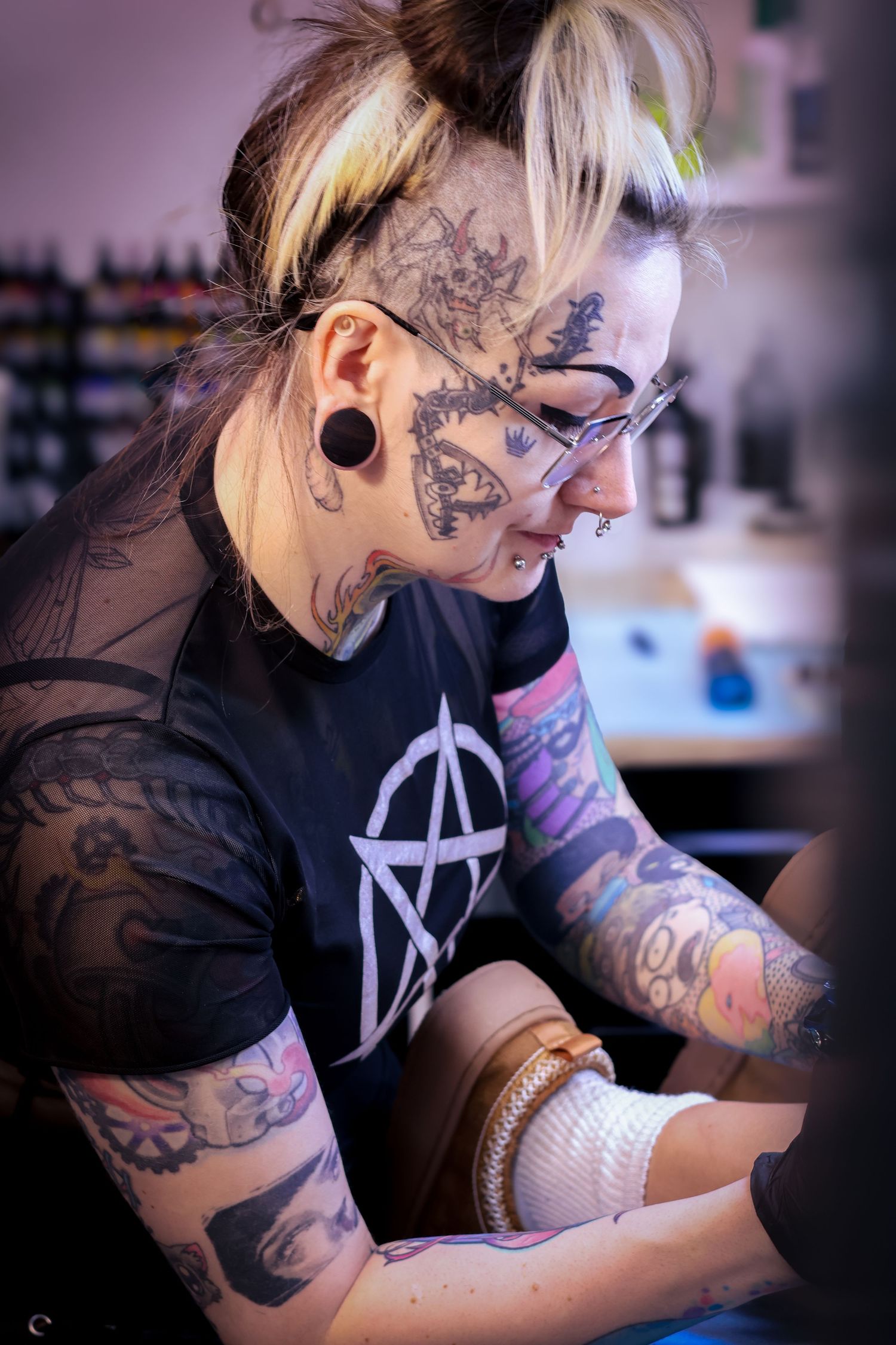 Tattoo artist with blonde hair and glasses works meticulously on a detailed design in a studio setting.