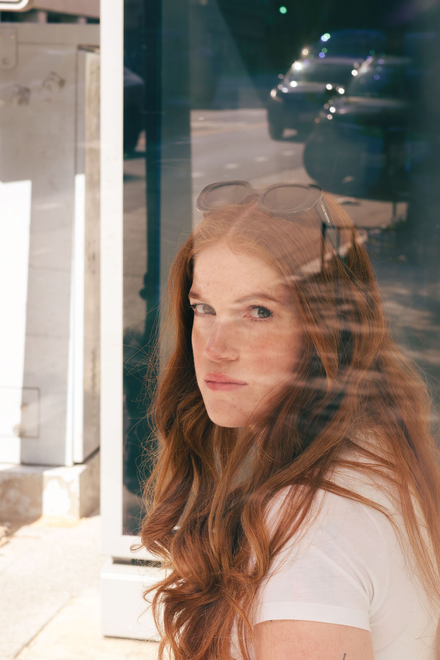 Close-up of freckled redhead woman with sunglasses on head near reflective glass window.