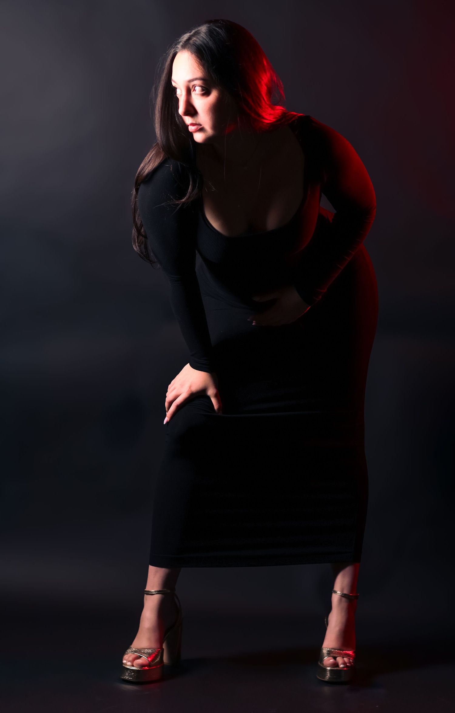 Woman in black long-sleeve dress posing dramatically with red lighting against dark background.