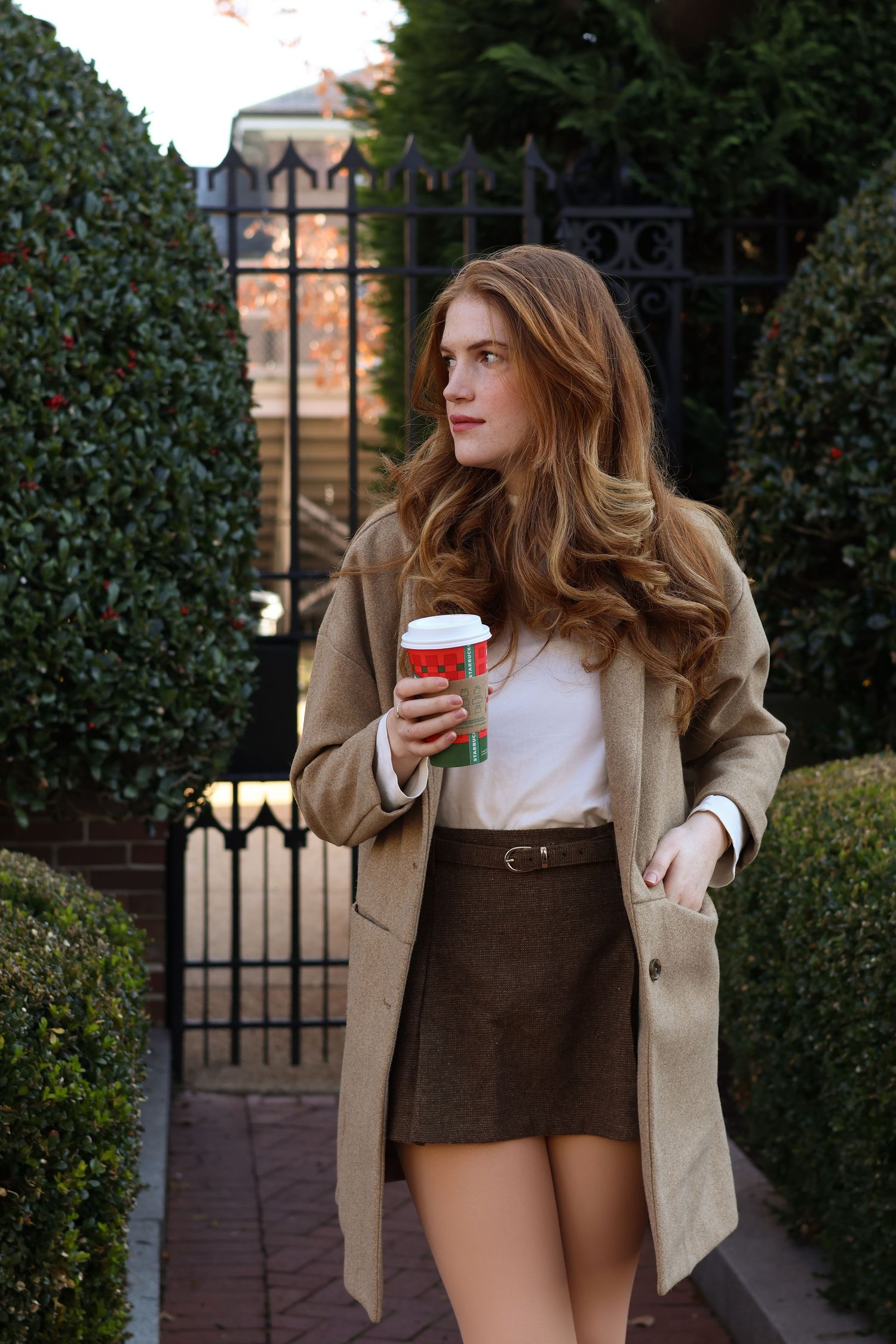 Stylish woman with long wavy hair wearing a camel coat and olive mini skirt, holding a festive red cup near iron gates.