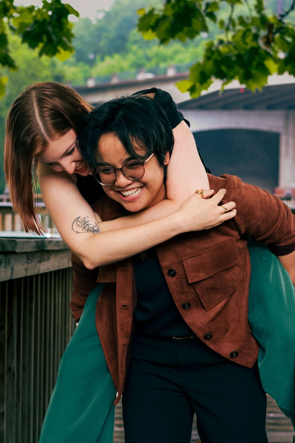 A smiling person with short dark hair and glasses in a brown jacket gives a piggyback ride to a woman hugging them under leafy trees.