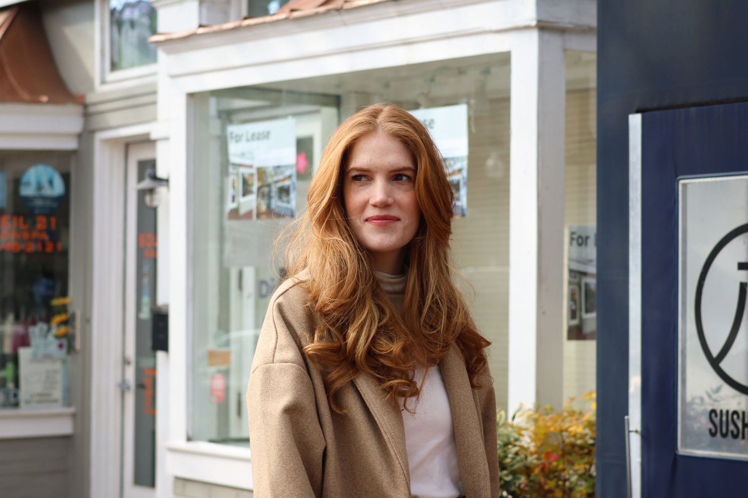 Young woman with long red hair wearing a beige coat standing on a European city street.
