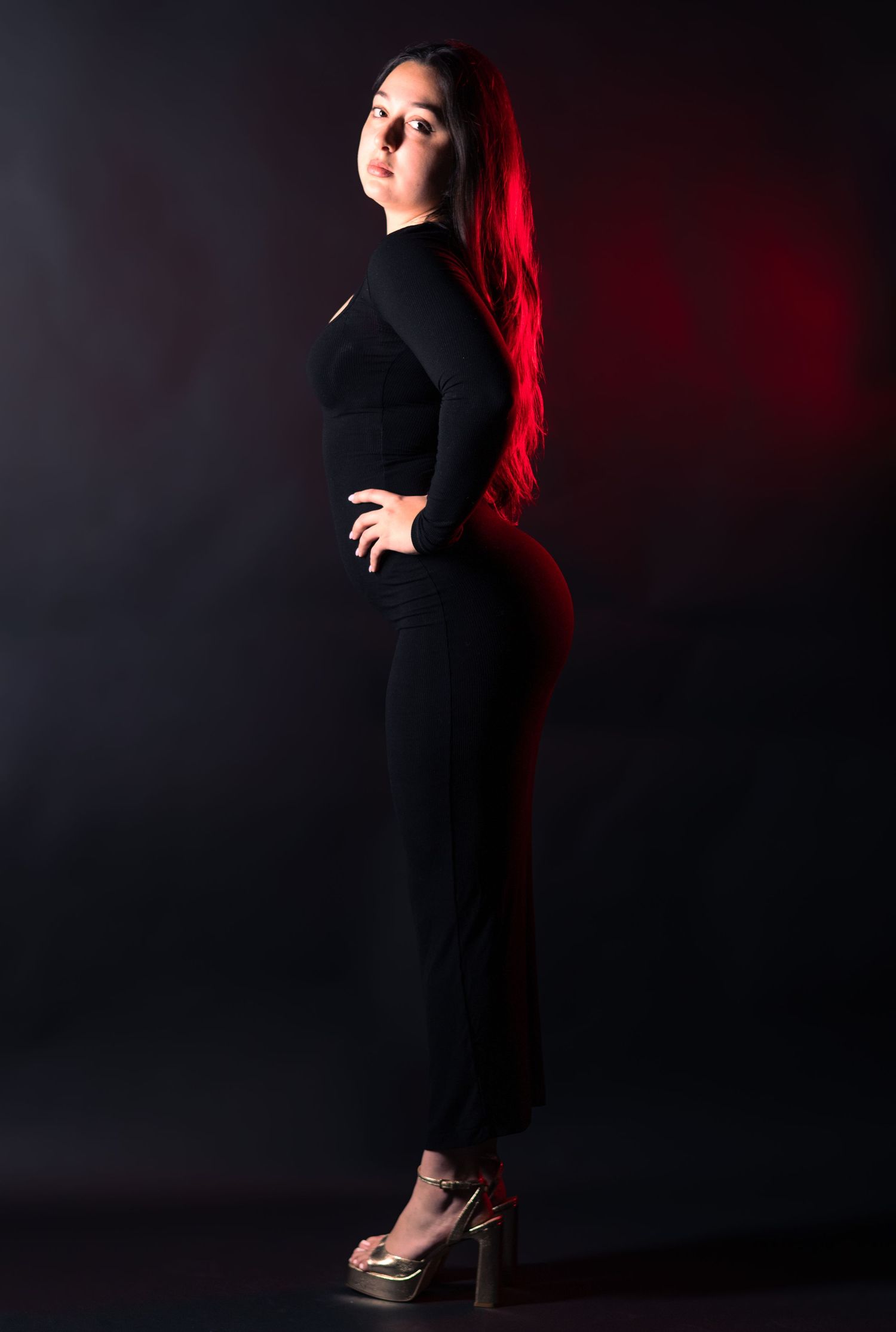 Woman with red hair in black outfit posing sideways with dramatic red studio lighting.
