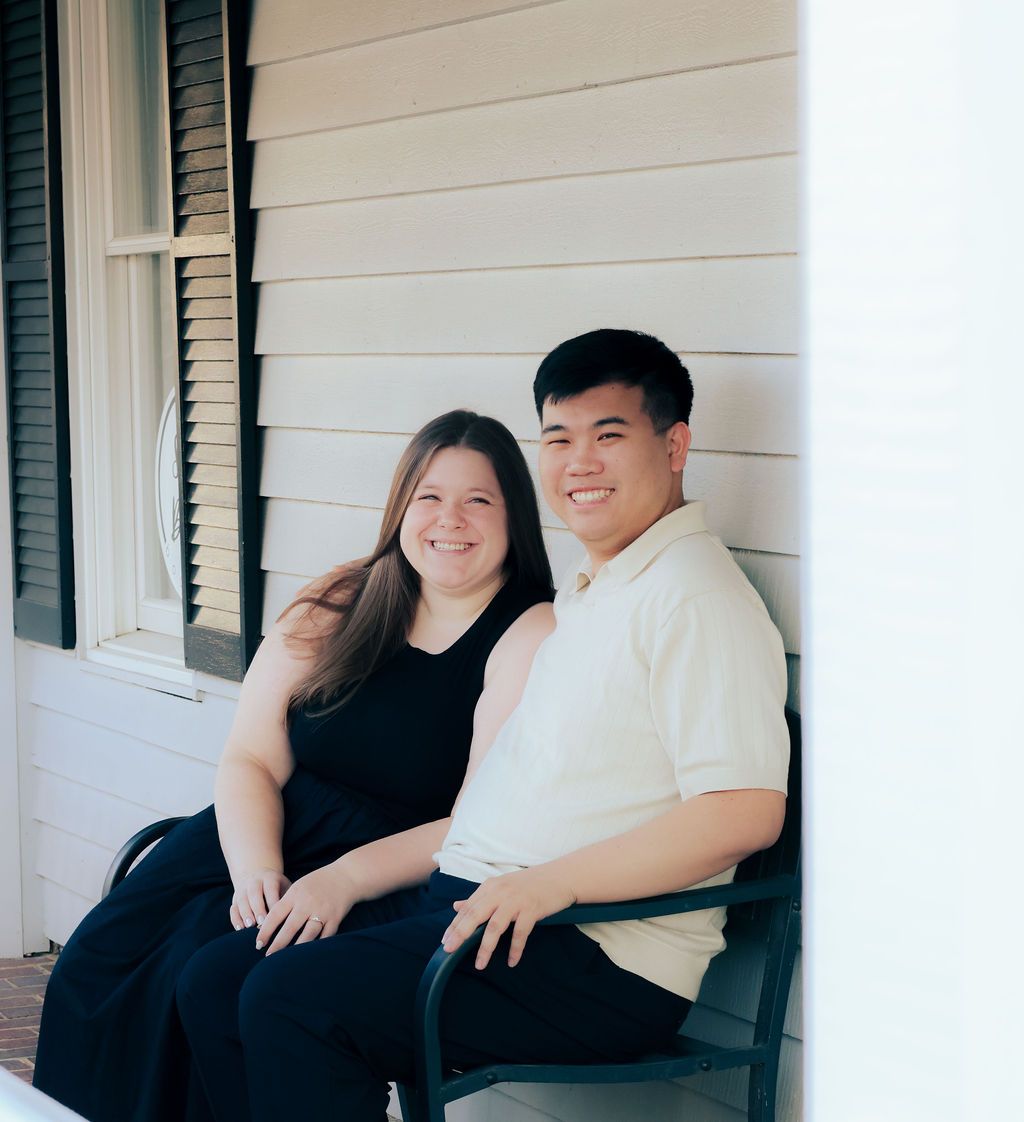 Smiling couple sitting side by side on a front porch bench beside a white sided house with dark window shutters.