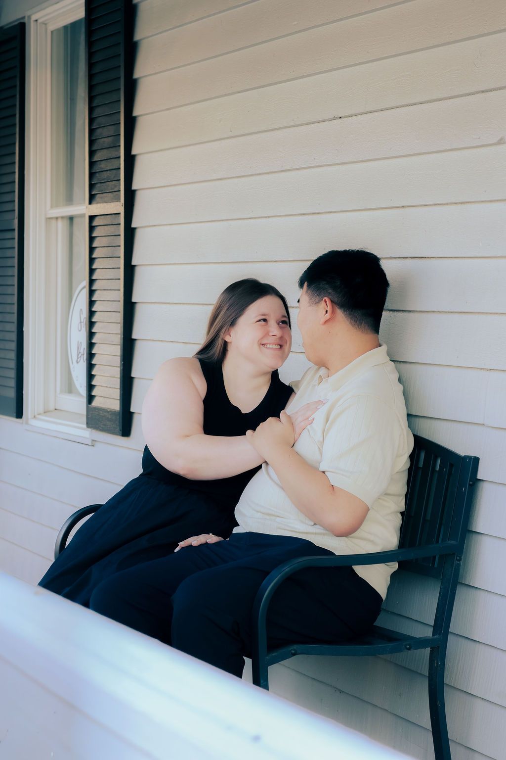 Smiling couple sitting close together on a black bench on a white-sided porch, embracing in a warm, affectionate moment.