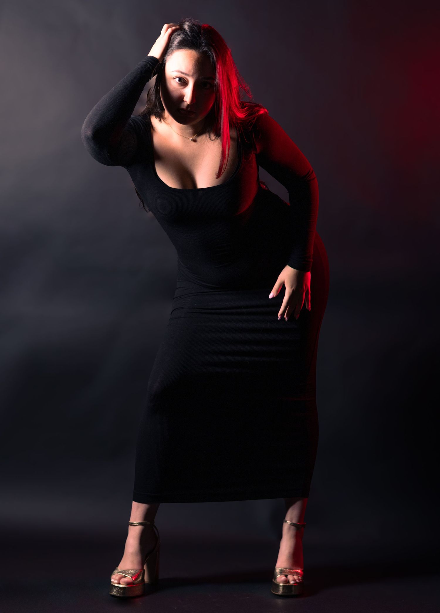 Woman in black form-fitting dress with red lighting posing dramatically against dark background.