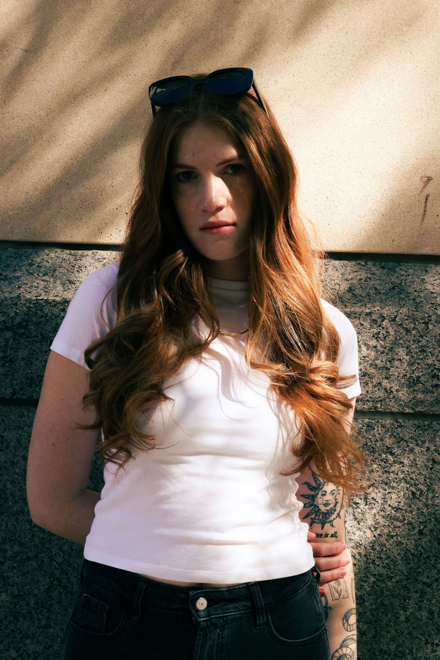 Young woman with long auburn hair and sunglasses on head, wearing white t-shirt and jeans, standing against a wall.