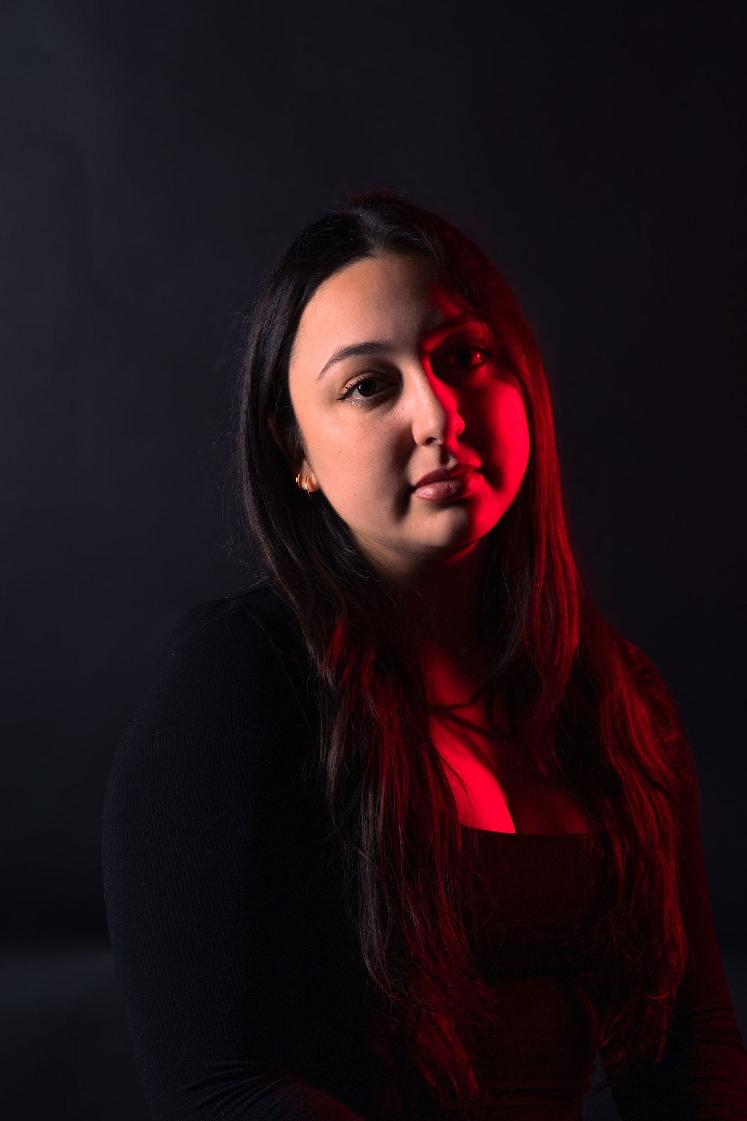 Young woman with long dark hair lit by dramatic red light against black background.