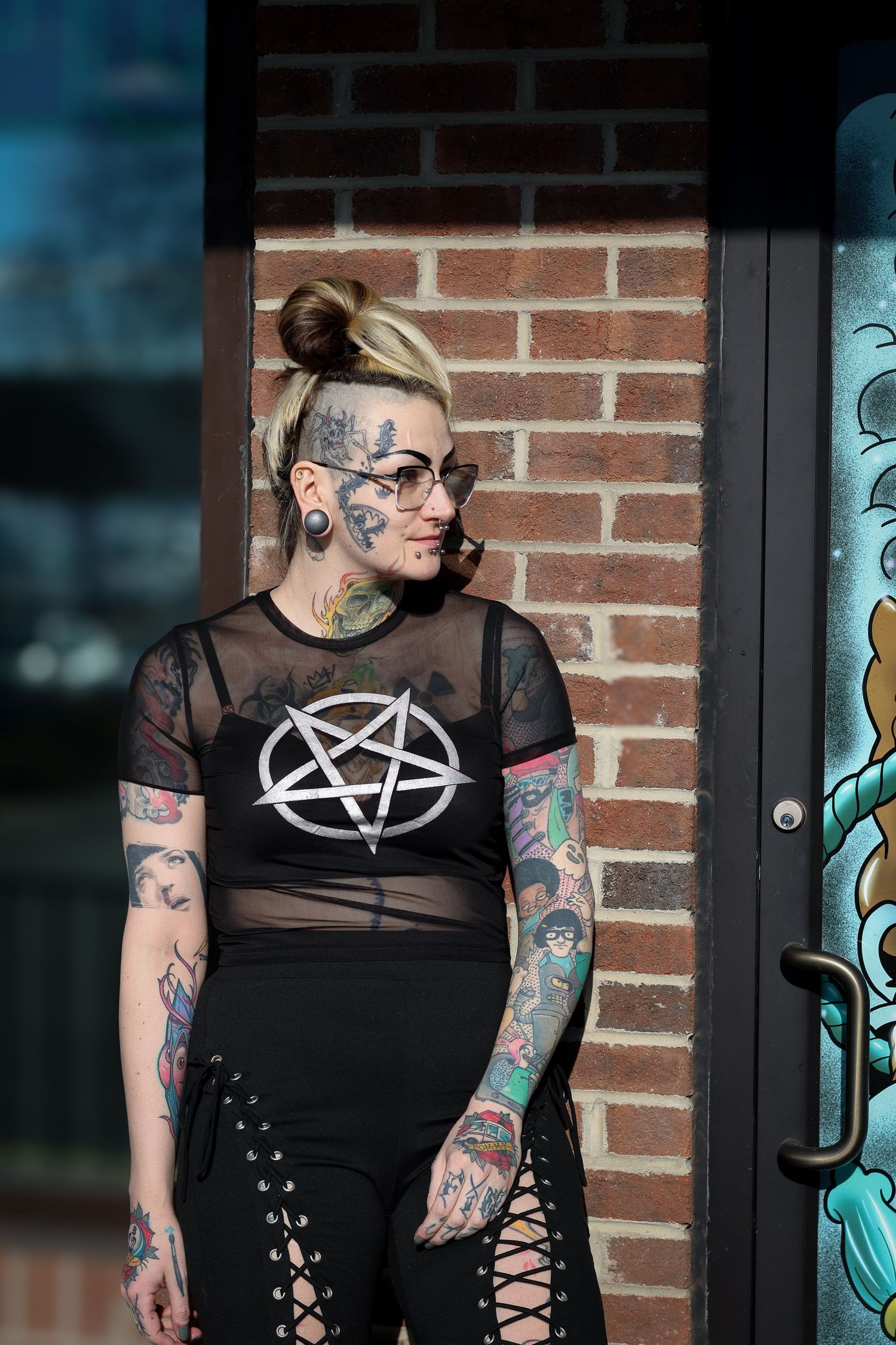 Person wearing black mesh top with tattoos and piercings stands against brick wall with turquoise artwork.