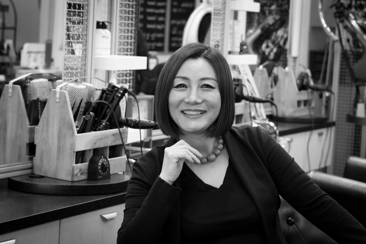 Black and white portrait of someone sitting at a counter with hand resting under chin, smiling warmly at camera.