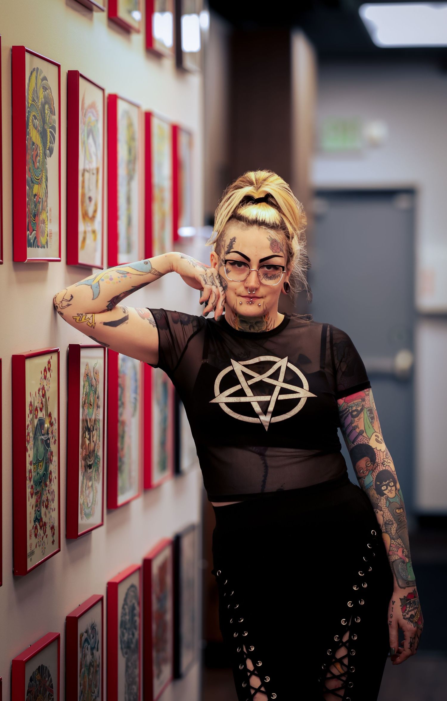 A person wearing glasses and a black t-shirt with tattoos poses against a wall with framed artwork.
