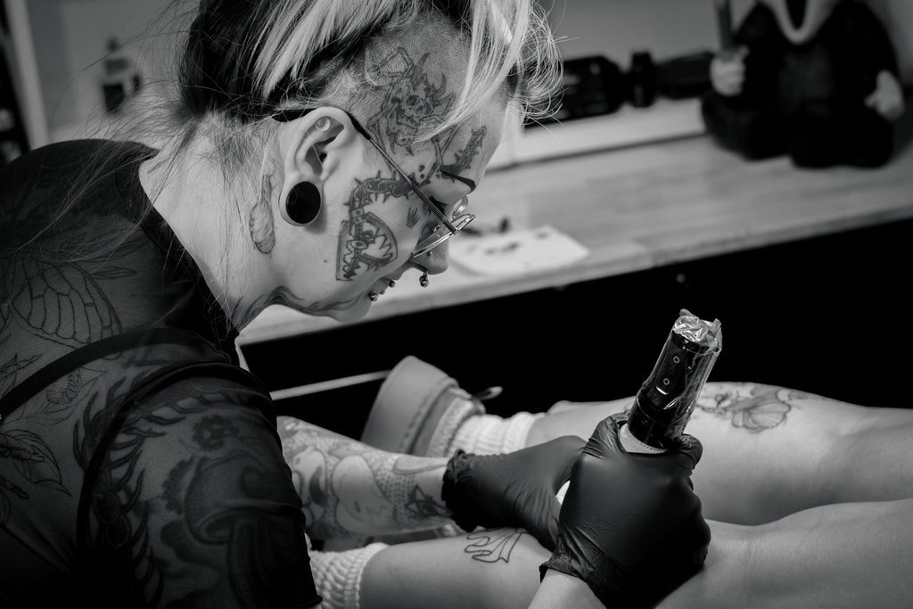 Tattoo artist with facial tattoos and gauged ears wearing black gloves and glasses, tattooing a client's leg with a tattoo machine.