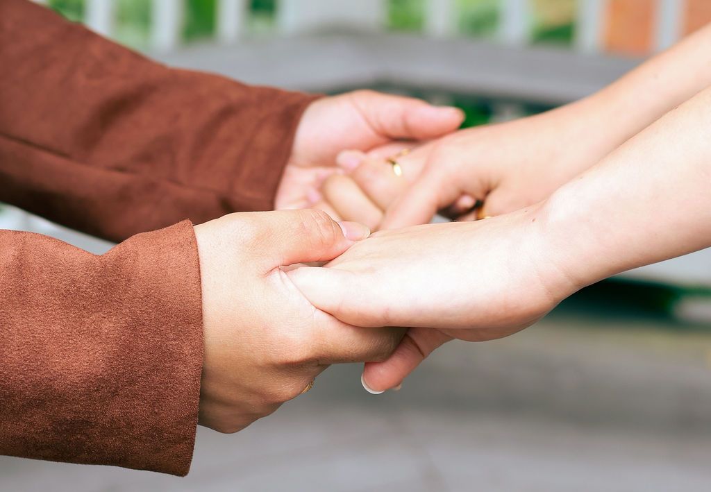 Two people in warm brown sleeves holding hands in a comforting, supportive gesture against a softly blurred outdoor background.