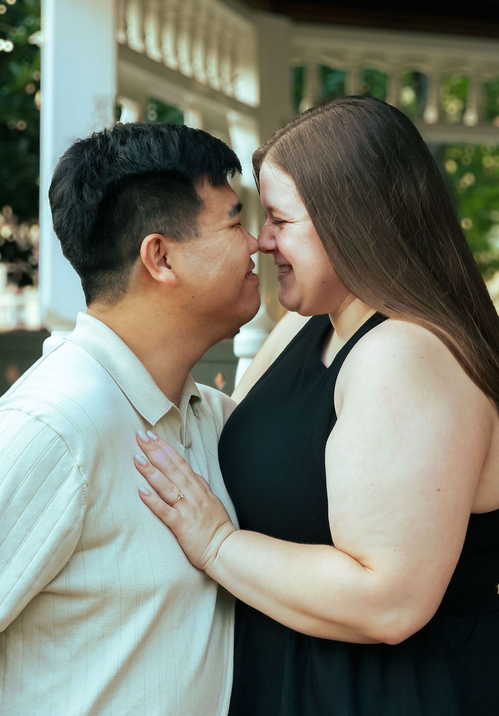 Smiling couple touching noses in a tender embrace outdoors, with the woman's hand resting on his chest near a white gazebo.