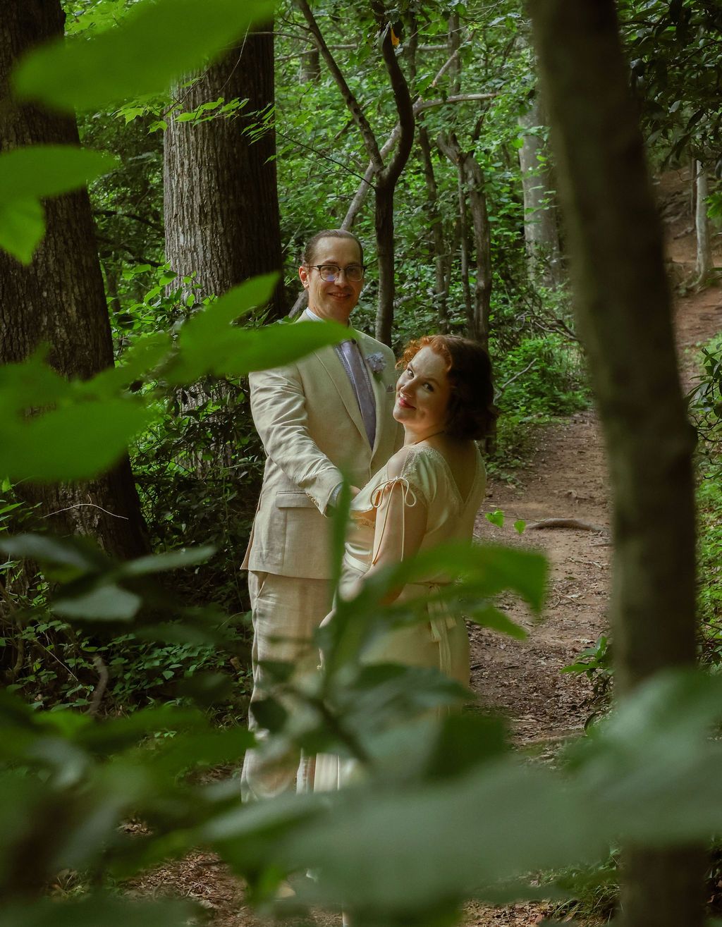 Couple in cream-colored outfits embracing on a wooded forest path, framed by green leaves and tall trees.