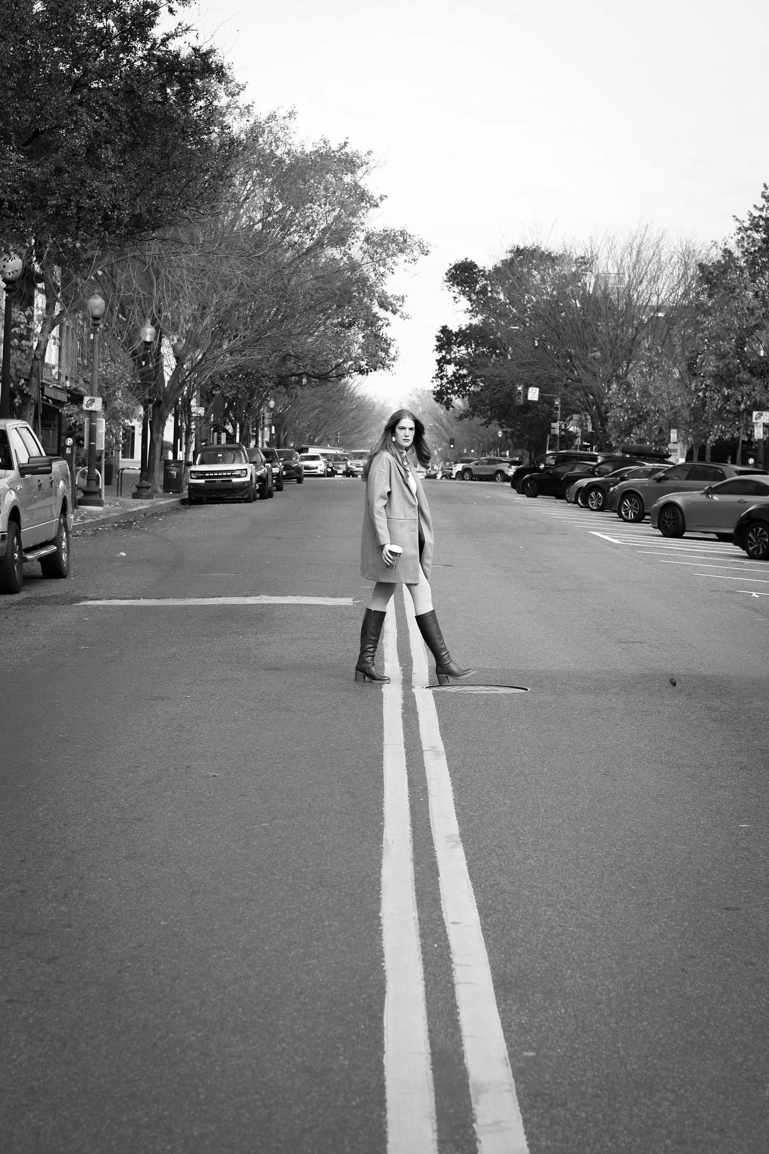 Woman in long coat and knee-high boots stands on urban street centerline in black and white photo.