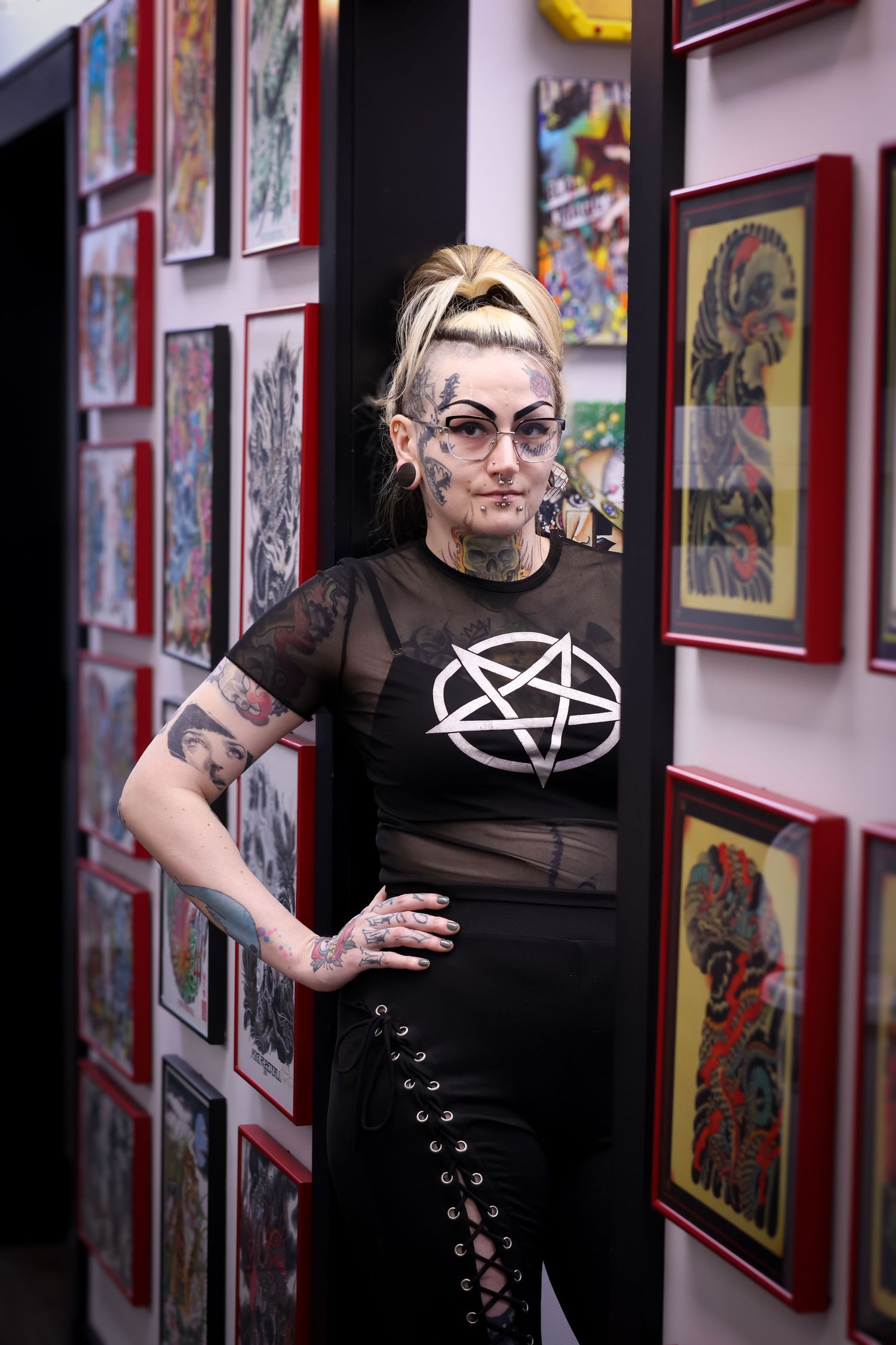 A person in black clothing and glasses poses in a gallery space with framed artwork on red walls.