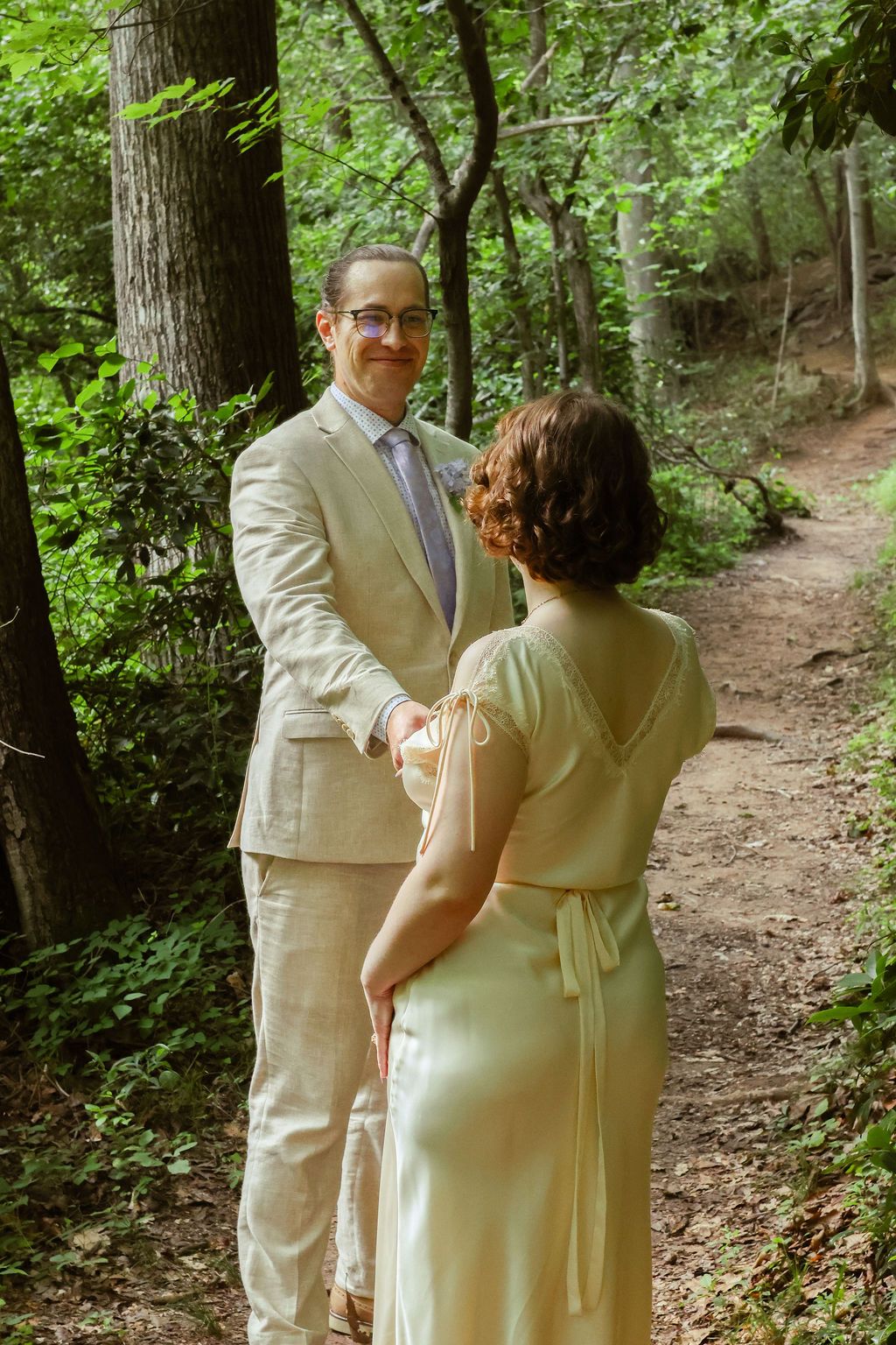 A man in a tan linen suit and glasses smiles and holds hands with a woman in a pale yellow dress on a wooded forest trail.