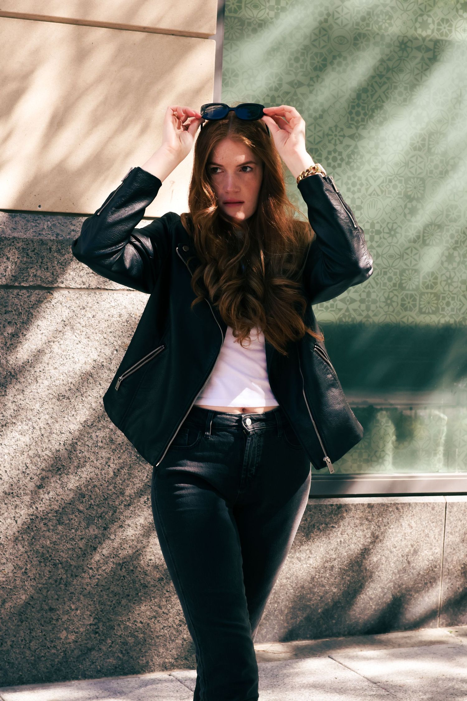 Young woman in black leather jacket, white tee, and black pants poses outdoors, lifting sunglasses to her head.