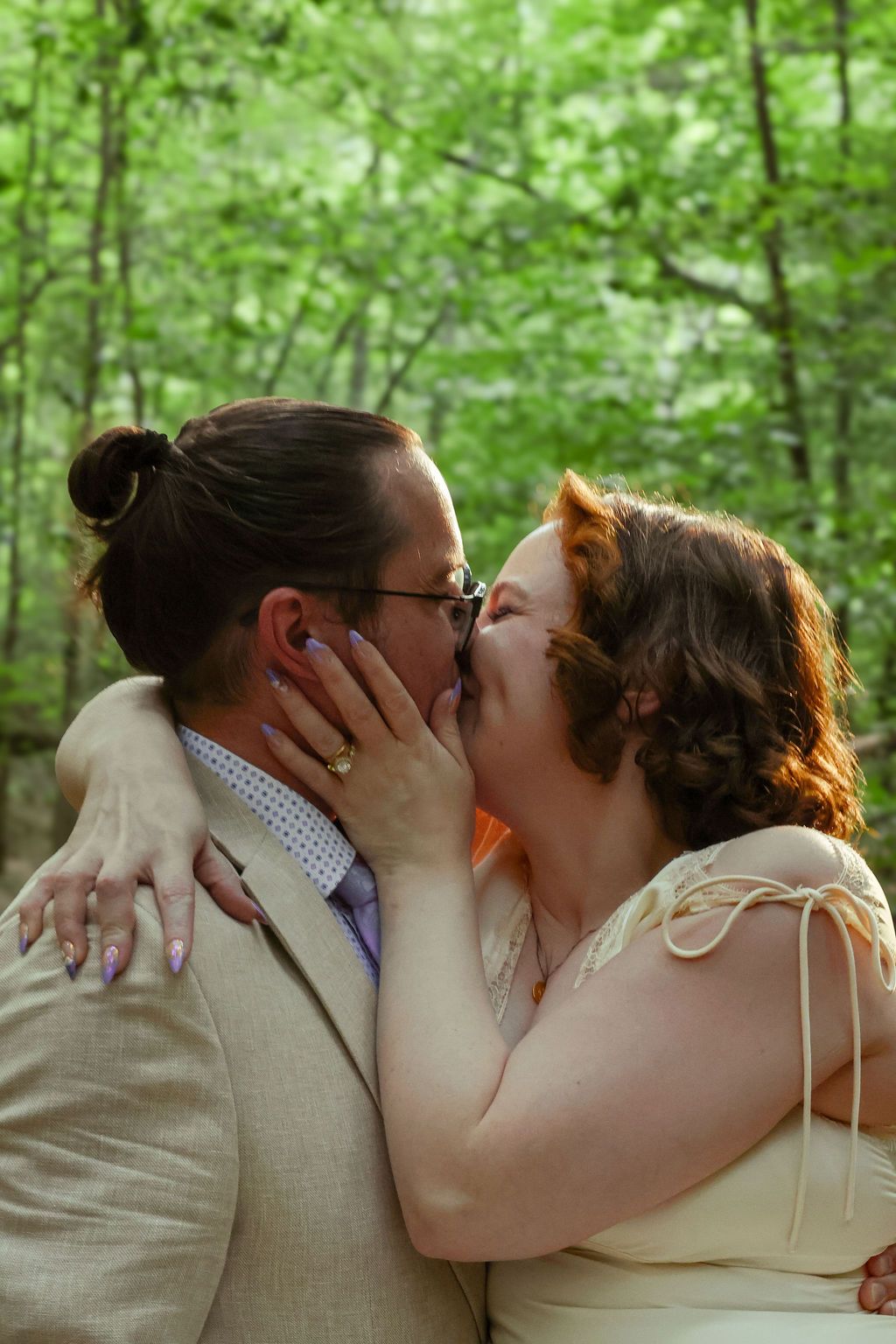 A couple in wedding attire shares a kiss in a lush green forest, her hand with painted nails and a ring on his cheek.