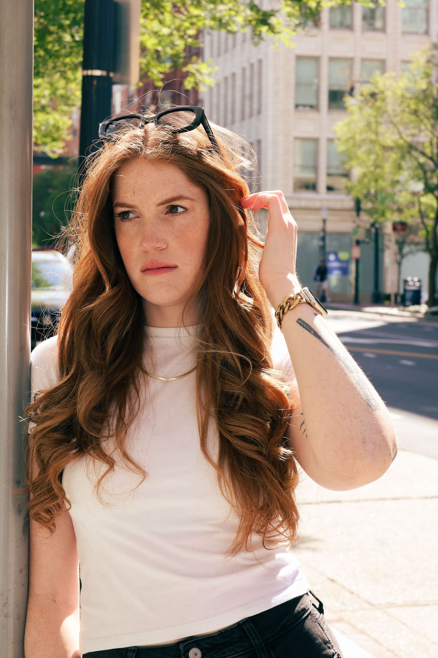 Young redheaded woman in white t-shirt adjusting sunglasses on head while leaning against urban building.