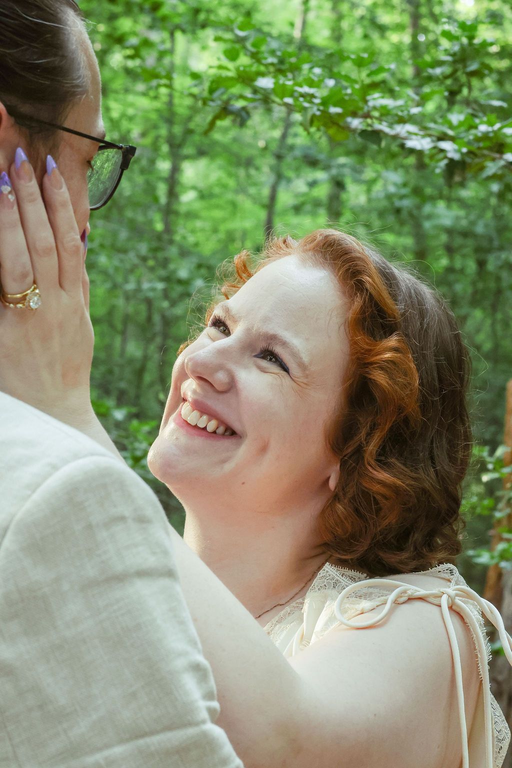 A smiling woman with curly red hair gazes lovingly as her partner gently touches her cheek, with lush green trees in the background.