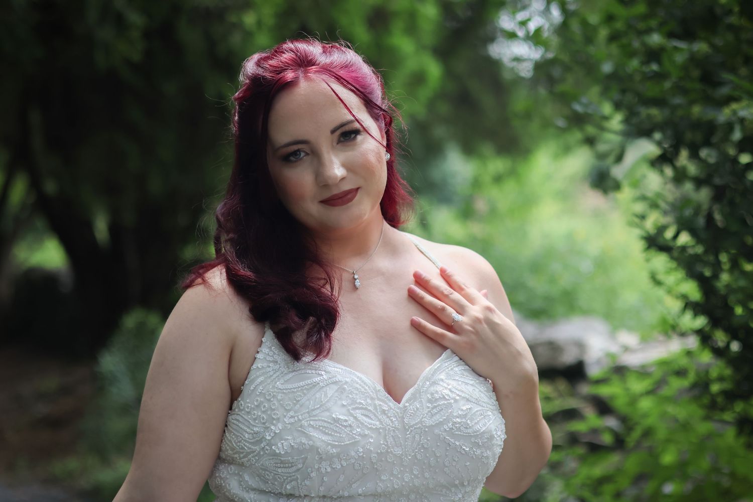 Beautiful bride with burgundy hair poses in white lace wedding dress against lush green garden background.