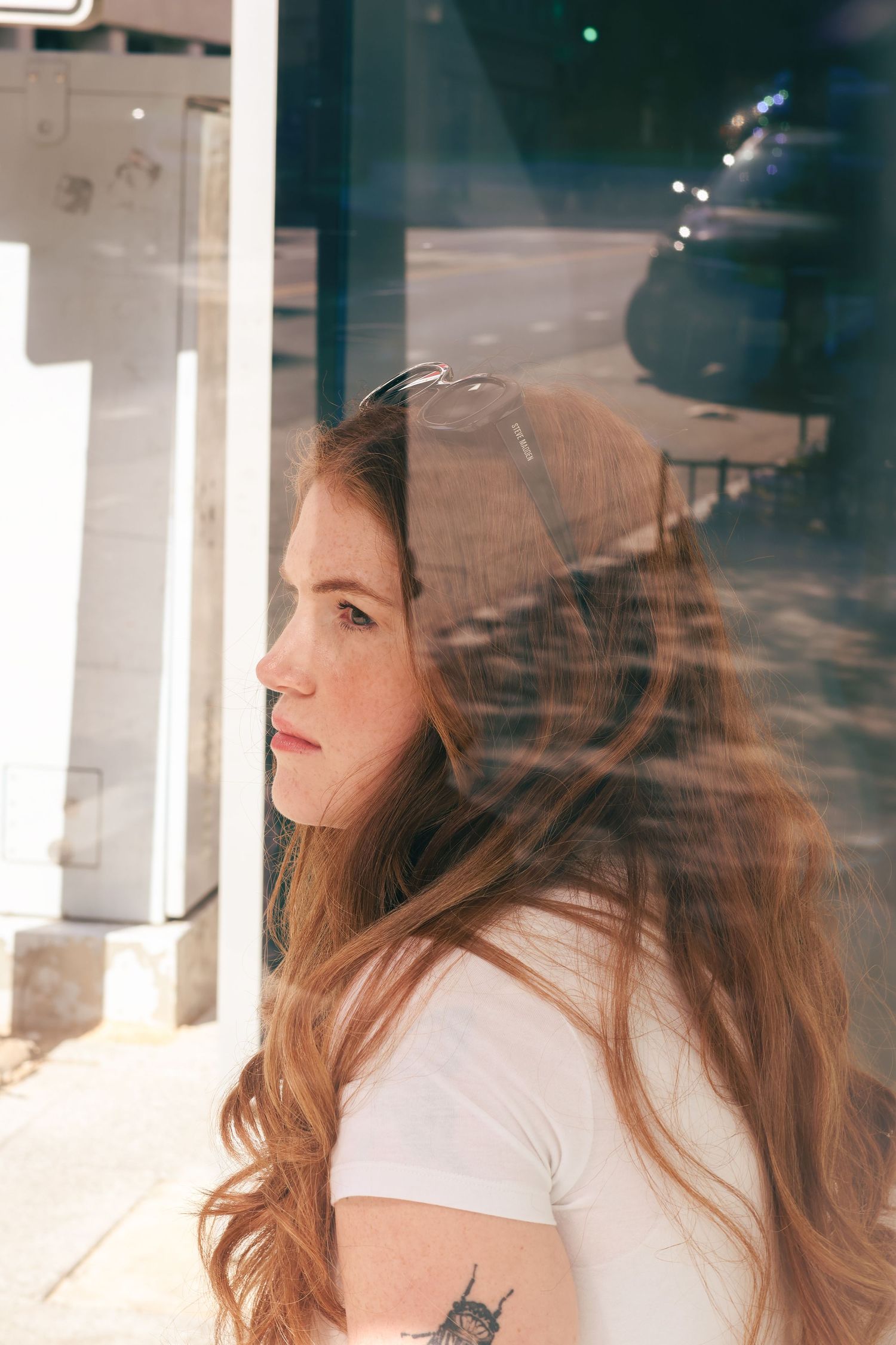 Side profile of young woman with flowing auburn hair leaning against glass window outdoors.