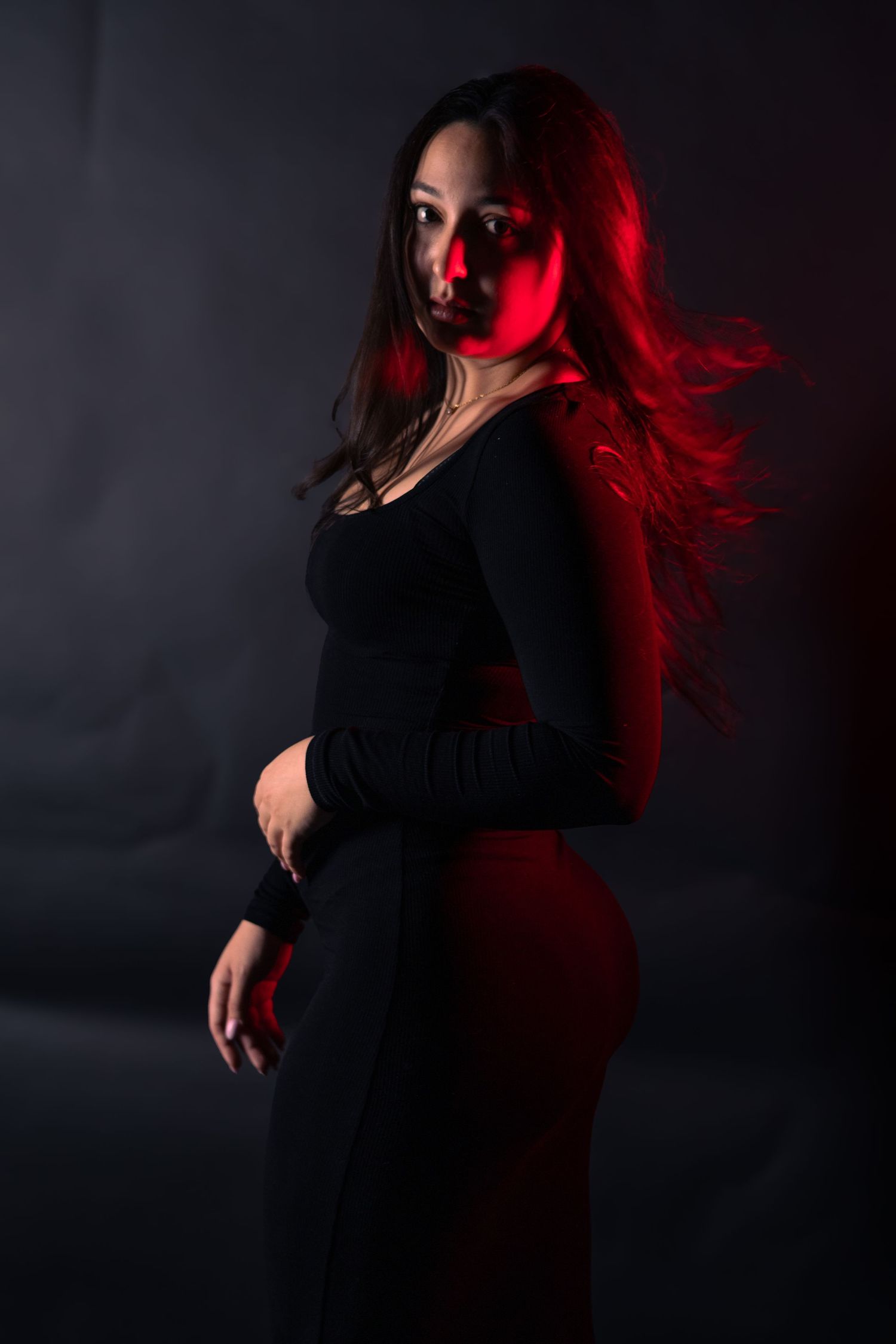 Woman in black outfit illuminated by red light posing against dark studio background.