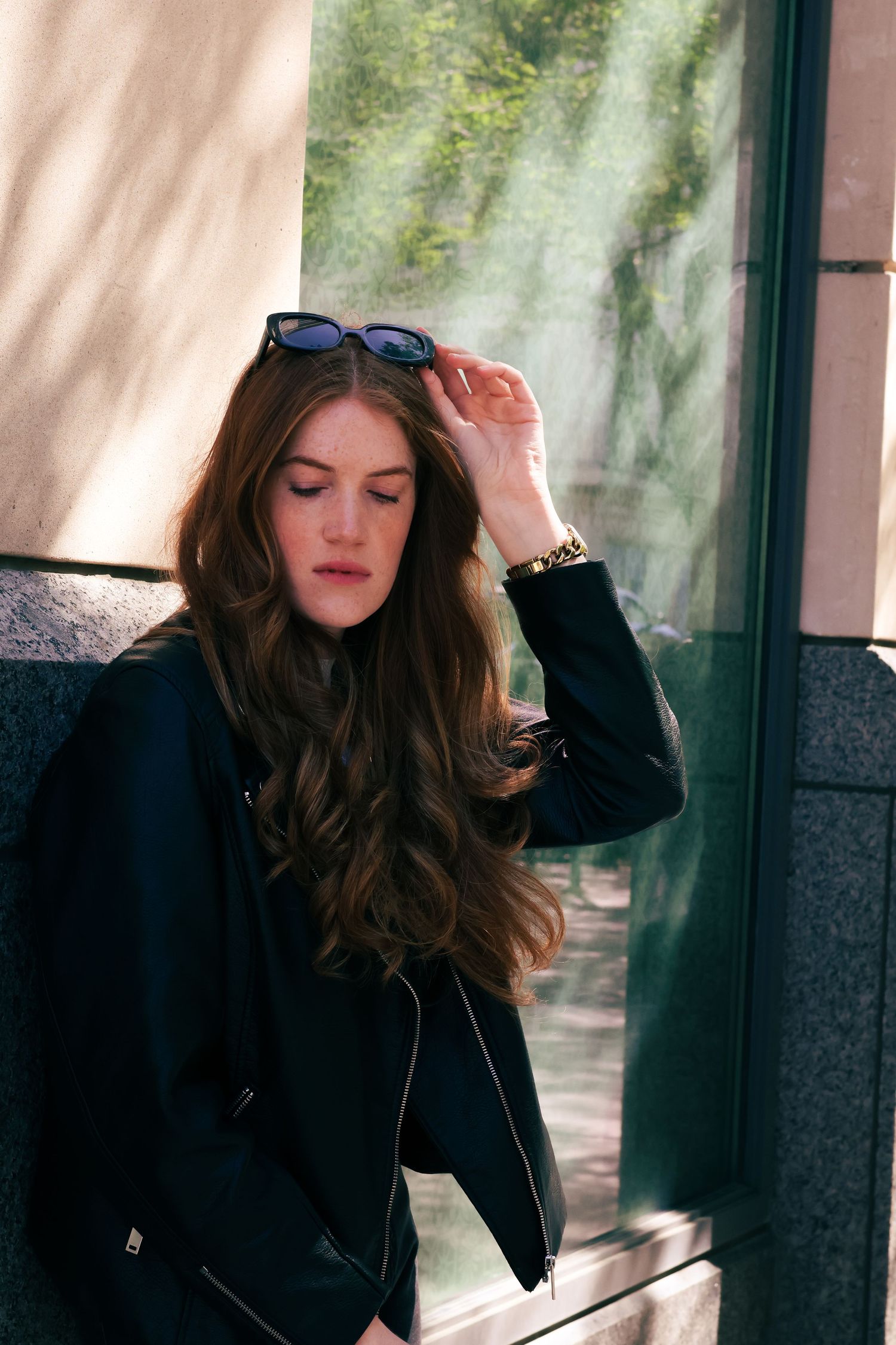 Young woman with long brown hair adjusting sunglasses wearing black leather jacket outdoors.