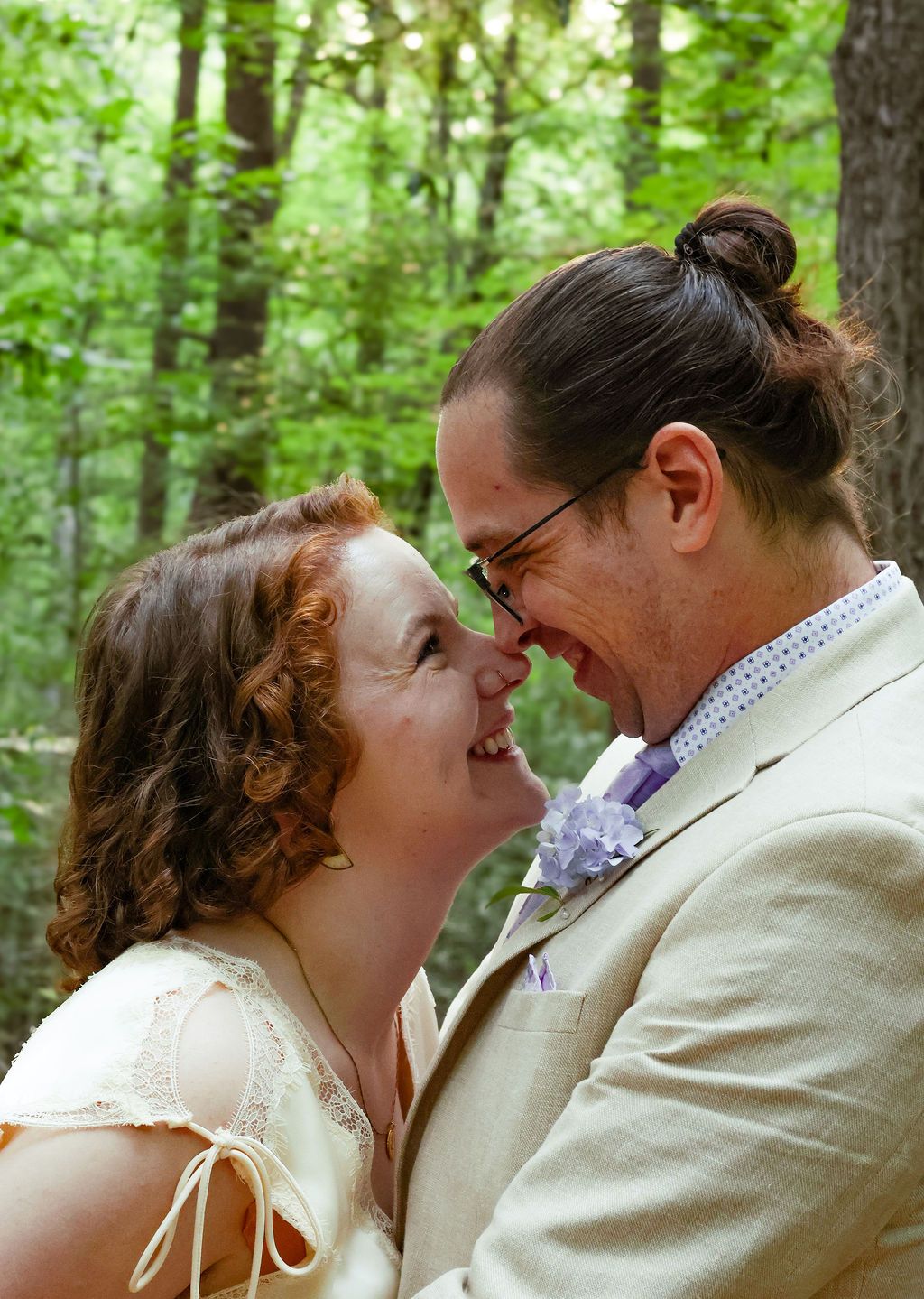 A smiling couple in wedding attire touch noses in a sunlit forest, the man wearing glasses and a lavender boutonniere.