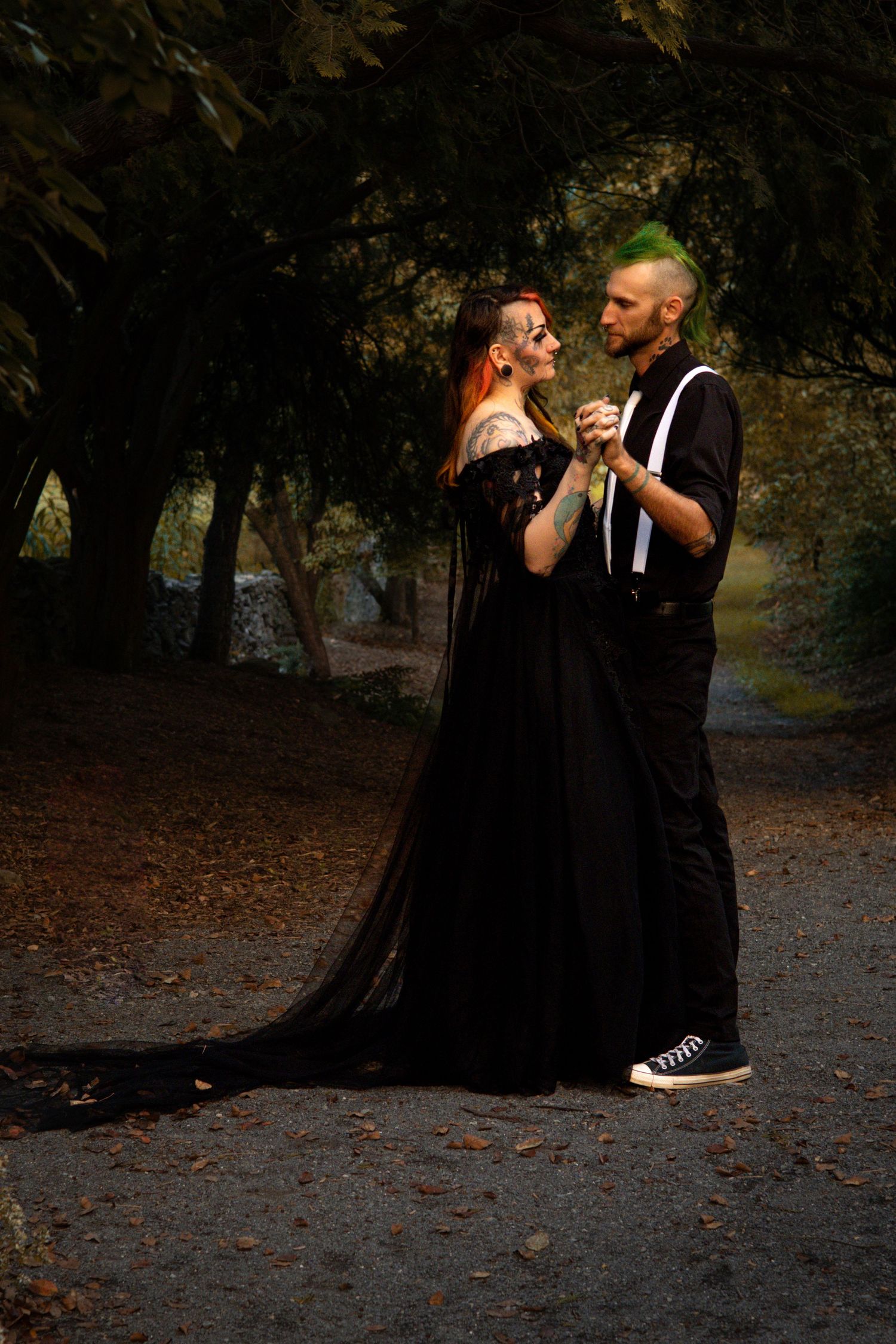 A couple in gothic-inspired black attire share an intimate moment while dancing on a dark forest path.