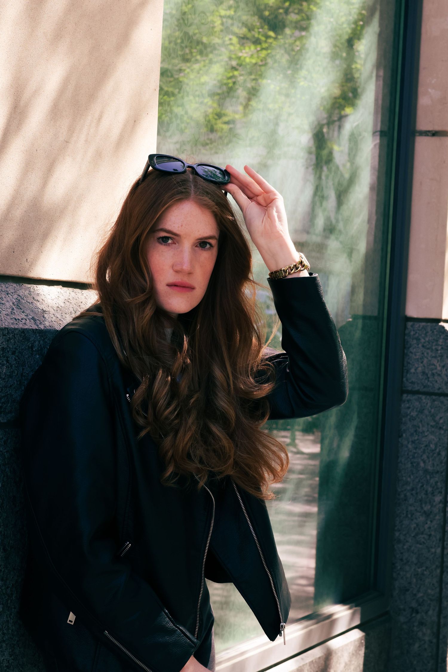 Stylish woman with long dark wavy hair holding sunglasses, wearing black leather jacket, leaning against wall outdoors.