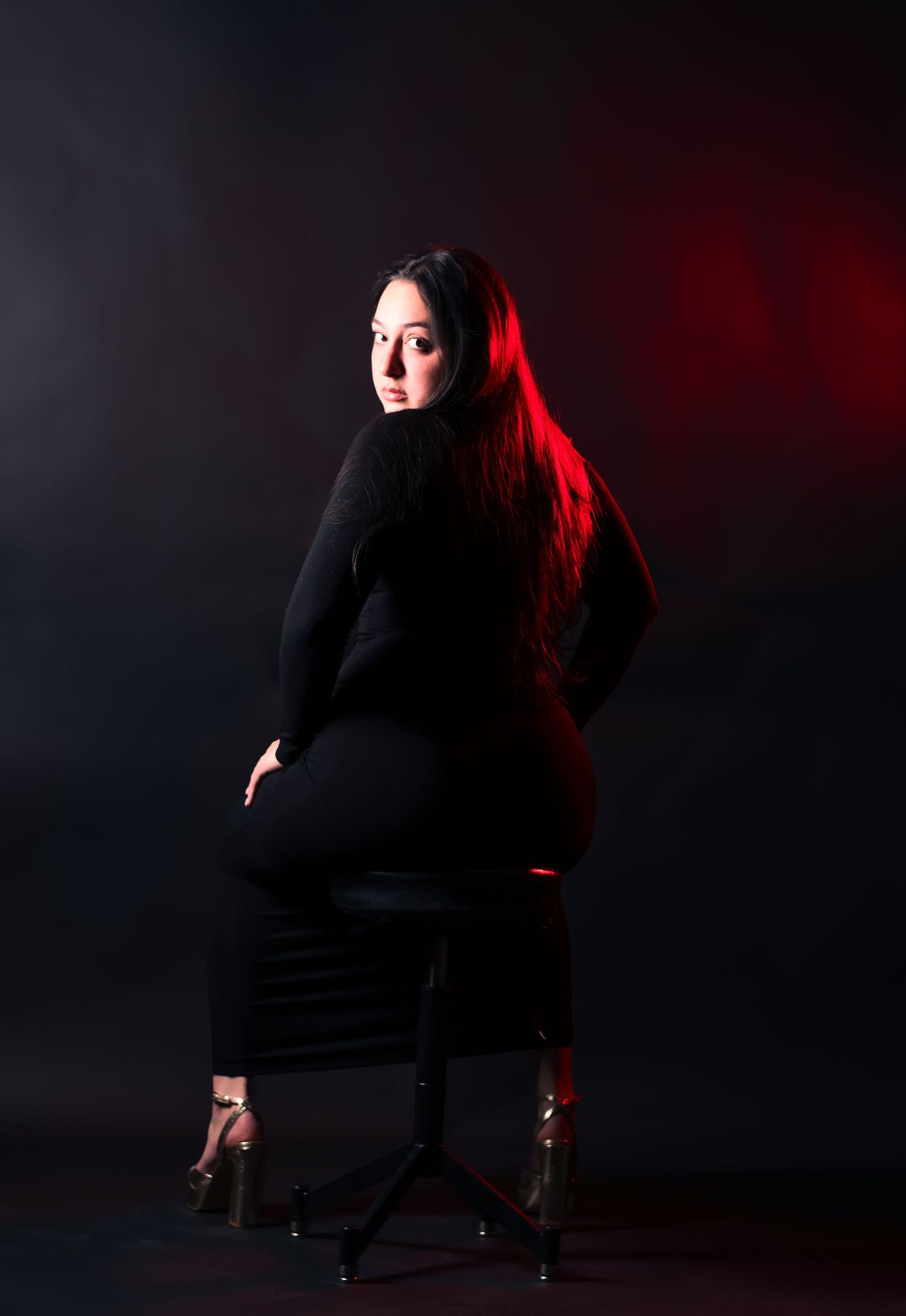 Woman in black outfit seated on stool with red lighting, looking back over shoulder.