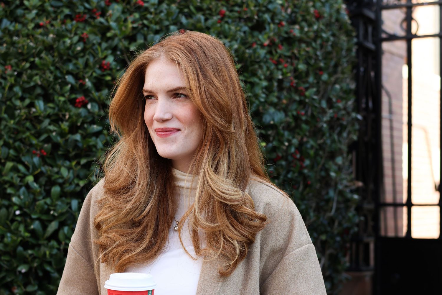Close-up of smiling redhead woman holding holiday coffee cup outdoors near green hedge in cozy beige sweater.