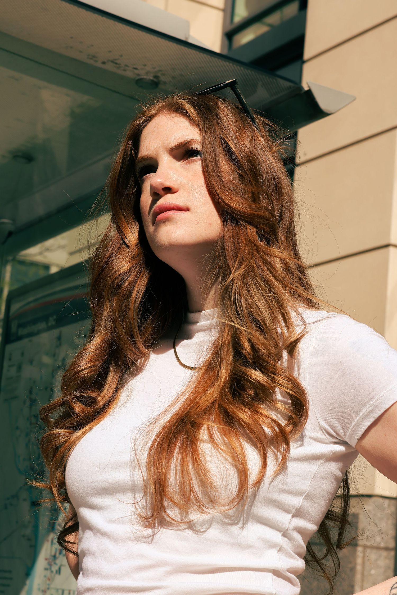 Young woman with long wavy auburn hair wearing a white t-shirt poses outdoors near a building in sunlight.