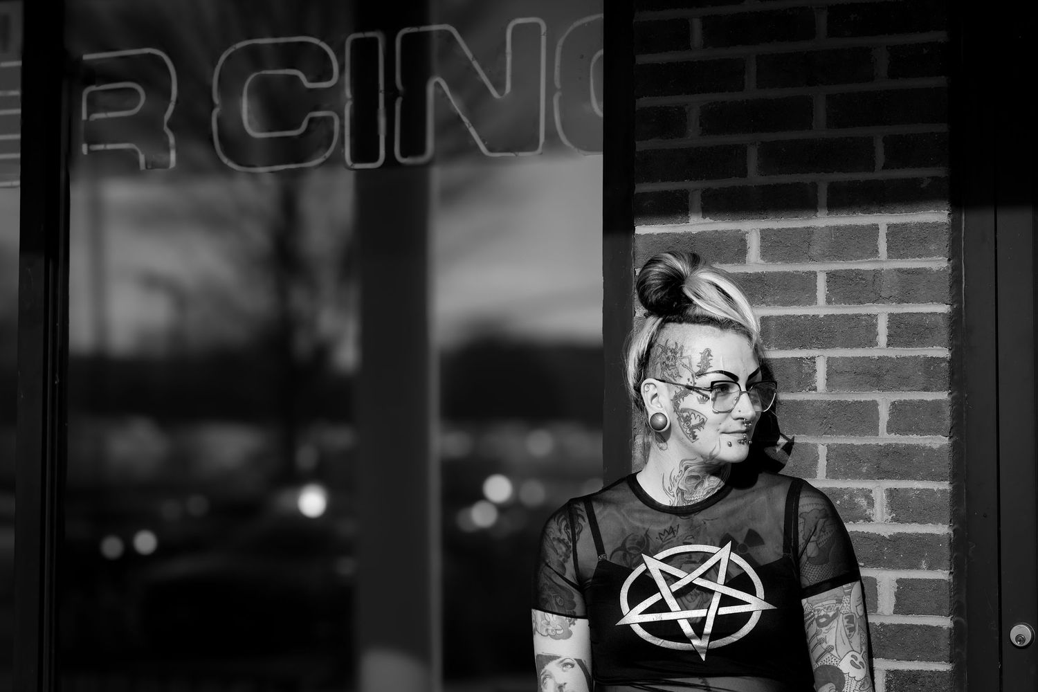 Black and white portrait of a person wearing glasses and a pentagram t-shirt standing against a brick wall.
