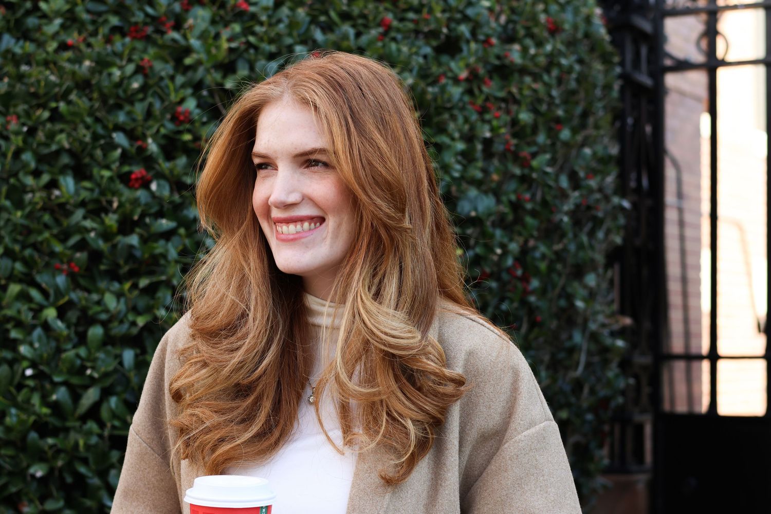 Smiling redhead woman in beige sweater holding a coffee cup outdoors near green hedges.