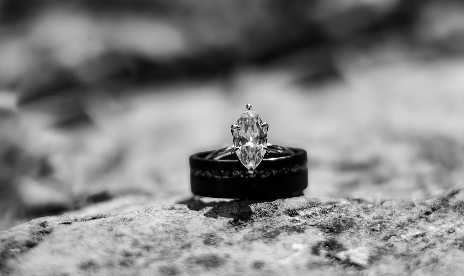 Black and white macro photograph of an elegant diamond engagement ring resting on a textured stone surface.