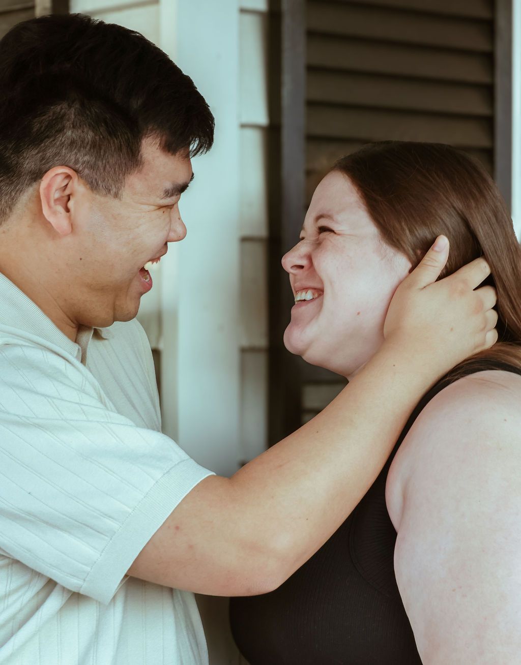 A smiling couple laughs together face to face as the woman gently holds her hair beside a white wall with shutters.