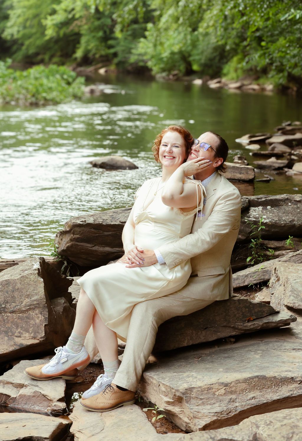 Joyful bride in a cream dress and groom in a matching suit laugh together on a rocky riverbank surrounded by green trees.