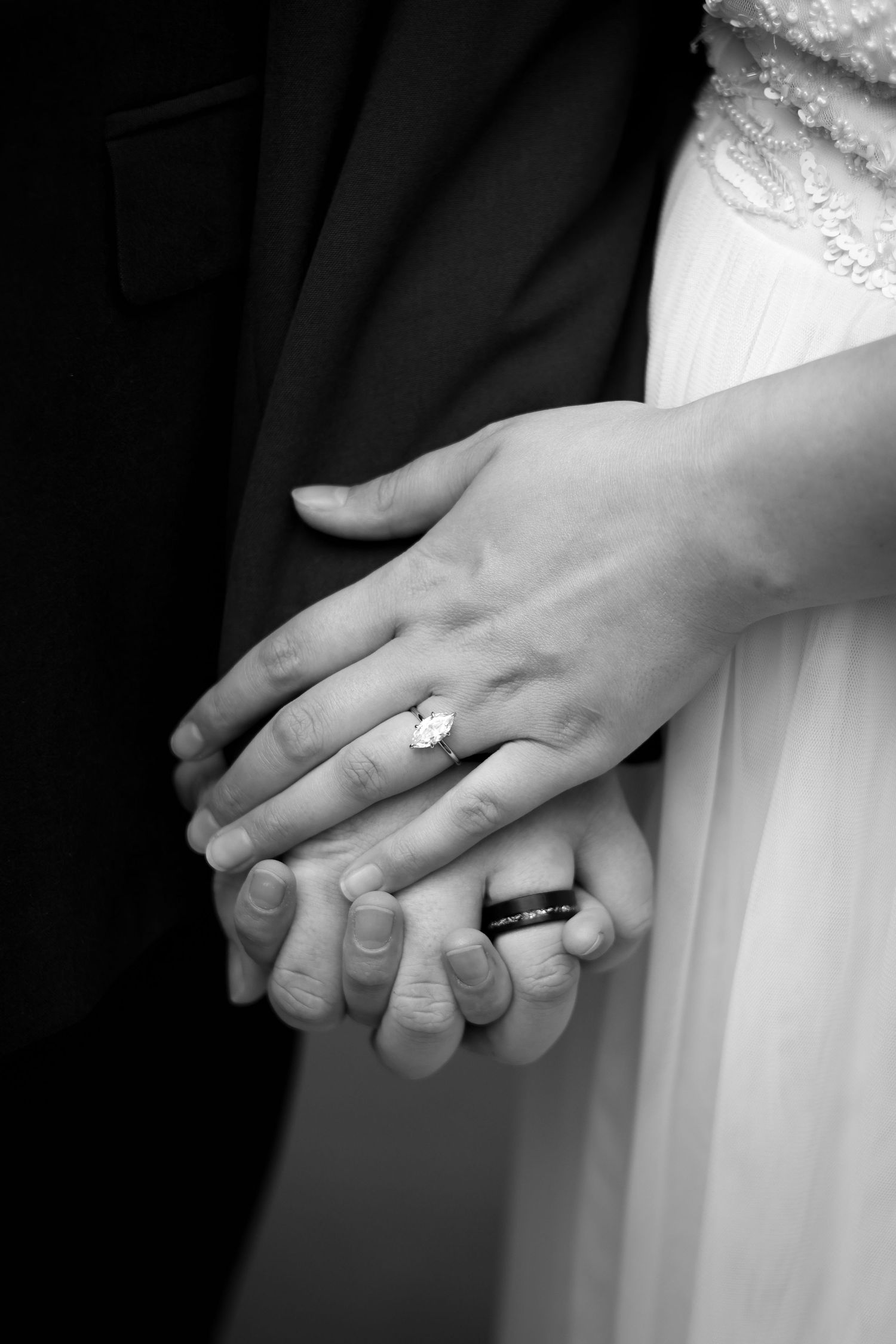 Close-up black and white photo of newlyweds holding hands showing wedding rings and formal attire.