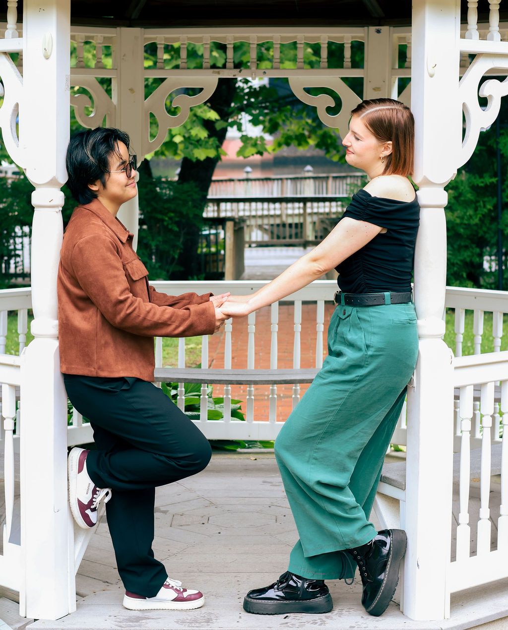 Two people hold hands while standing face to face in a white gazebo, one in a rust jacket and black pants, the other in green trousers.