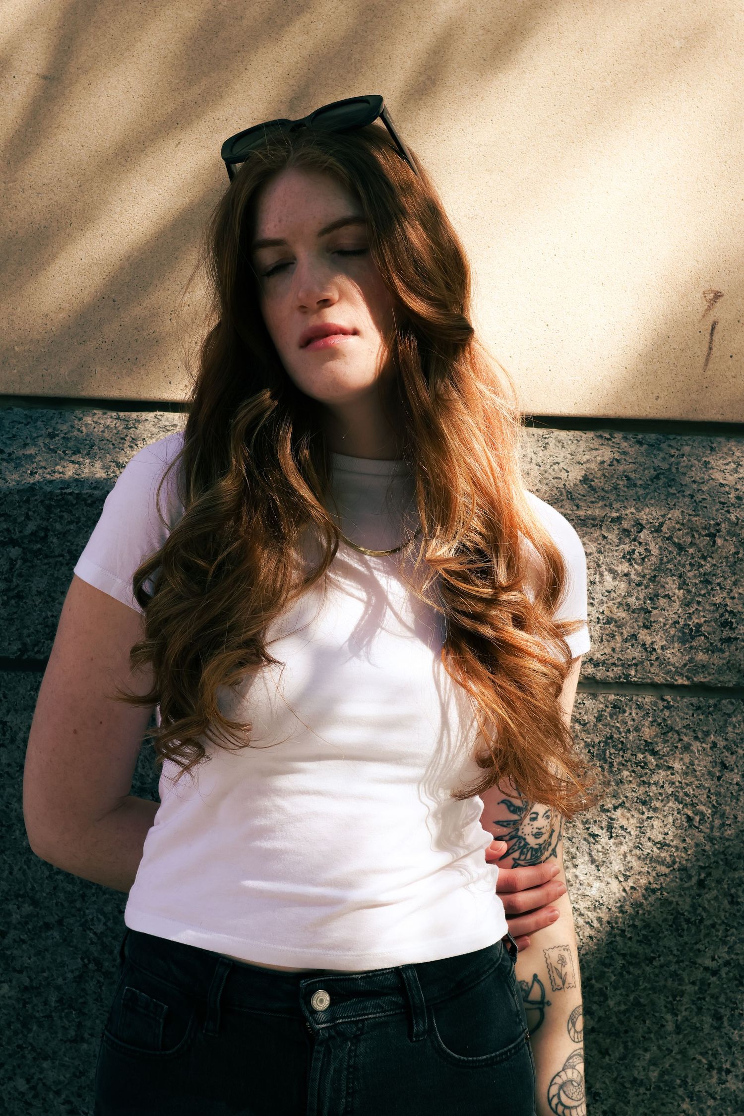 Young woman with long auburn hair and tattoos wearing a white t-shirt and sunglasses on head, posing outdoors.