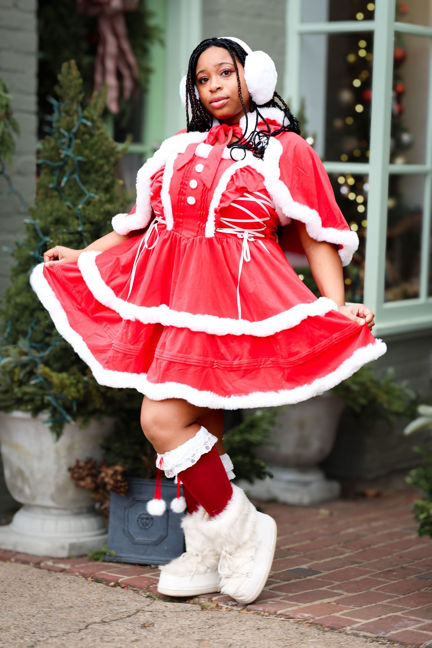 Woman in red Santa dress with white fur trim, earmuffs, and boots posing outdoors near Christmas decorations.