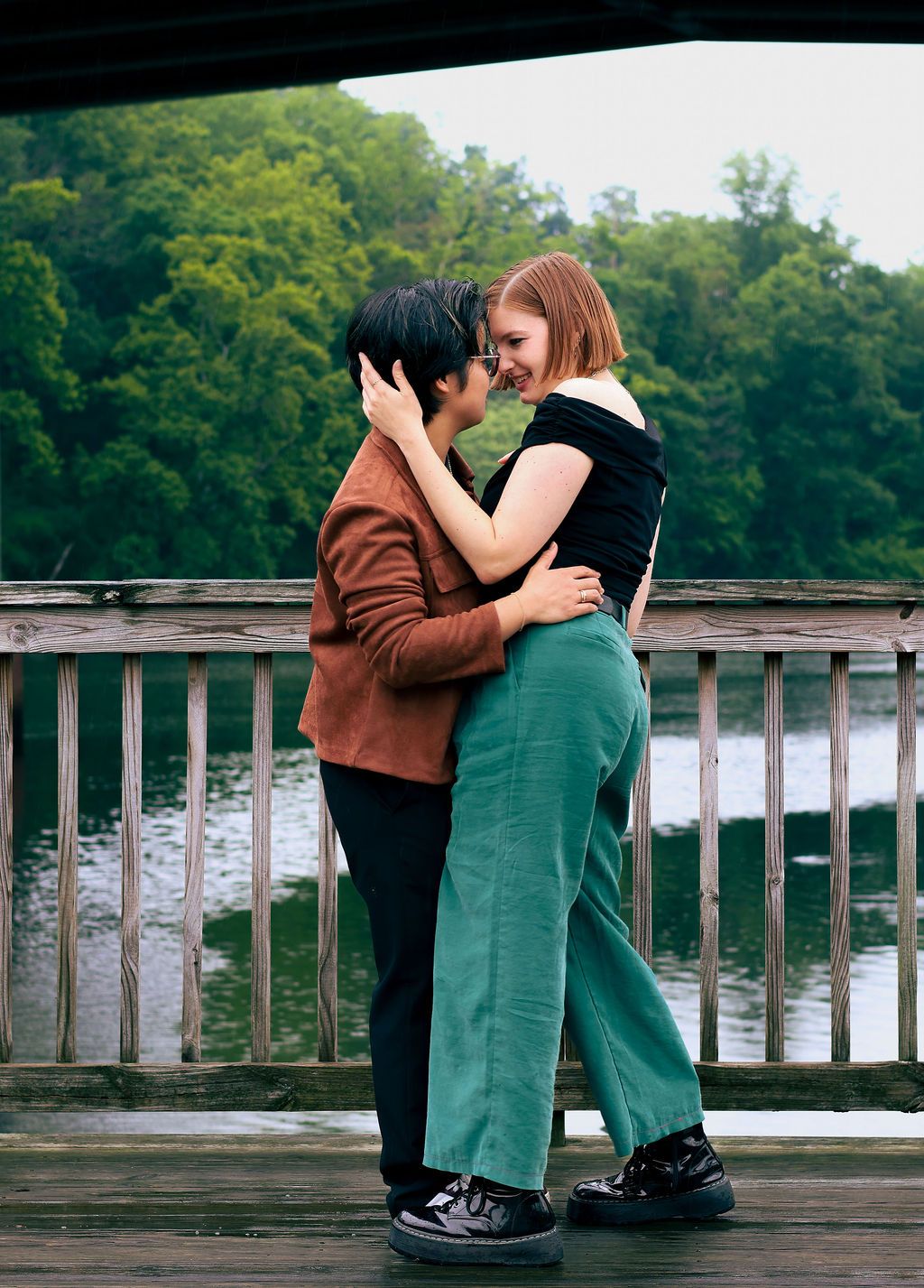 Two people in a tender embrace on a wooden lakeside dock, one in green pants and black top, with trees in the background.