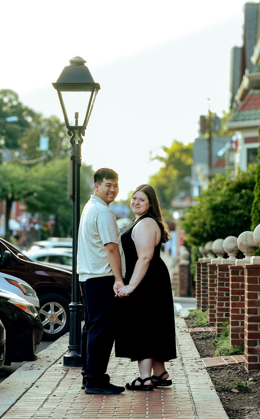 A couple holds hands and smiles on a brick sidewalk beside a vintage street lamp during an engagement photo session in a charming town.