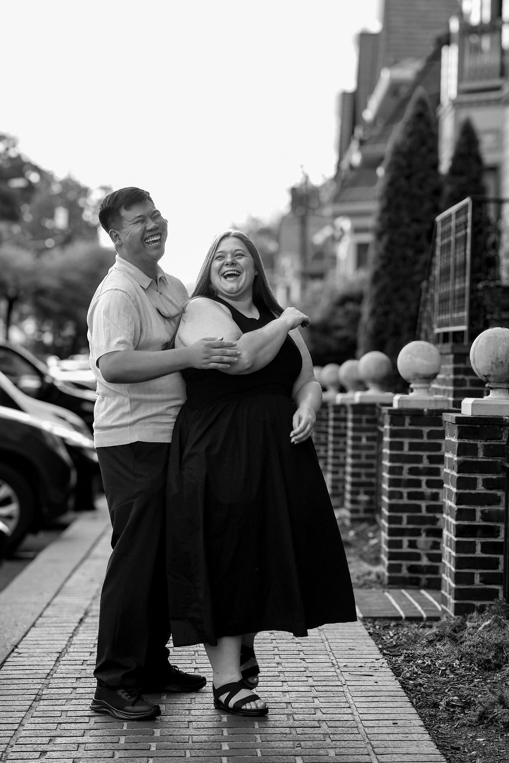Black and white photo of a laughing couple embracing on a brick sidewalk lined with brick pillars and parked cars on a city street.