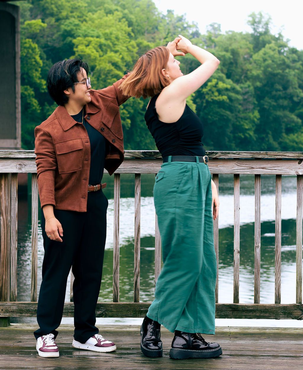 Two people pose on a wooden dock by a calm lake, one in a rust jacket and black pants, the other in a green skirt with arm raised.